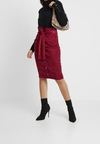 Burgundy mockaläder penningkjol med frontknut, knappar fram, kombinerad med en svart långärmad topp och svarta klackstövlar.