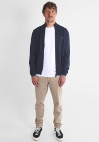 Veste zippée navy sur un T-shirt blanc, associée à un pantalon beige et des sneakers noires. Design simple avec des poignets élastiques et un détail de logo.