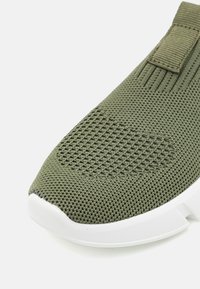 Oliwkowe buty sportowe z teksturowaną siateczką, gumową podeszwą i materiałową pętlą. Posiadają zaokrąglony czubek i wyściółkę amortyzującą.