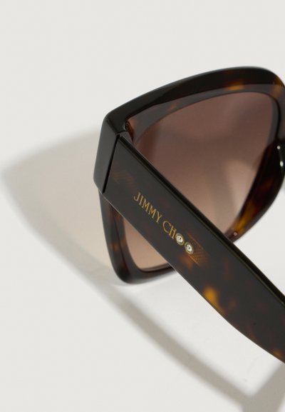 Lunettes de soleil en écaille de tortue avec une monture triangulaire, des verres dégradés marron, un logo "JIMMY CHOO" en doré et des accents décoratifs en pierre sur la branche.