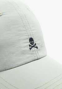 Light green cap made of fabric with contrasting navy stitching. Features a skull and crossbones logo embroidered on the front.
