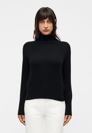 Jonge vrouw met donker haar draagt een zwarte coltrui en een witte broek, staand tegen een eenvoudige lichte achtergrond.