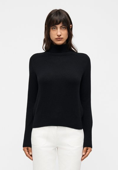 Jeune femme aux cheveux foncés portant un pull à col roulé noir et un pantalon blanc, debout devant un fond clair uni.