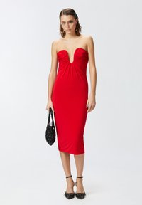 Crvena haljina bez naramenica s dubokim V-izrezom, nabiranjem na bustu i uskim krojem. Model drži crnu pletenu torbicu.
