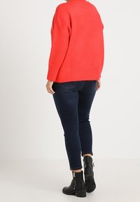 Roter Strickpullover mit lockerem Schnitt, kombiniert mit dunkelblauen Skinny-Jeans und schwarzen Ankle-Boots mit Reißverschluss und strukturierten Sohlen.