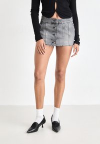 Jupe en denim grise courte avec un ourlet effiloché, dotée de boutons sur le devant et de coutures verticales, associée à des chaussures noires et des chaussettes blanches.