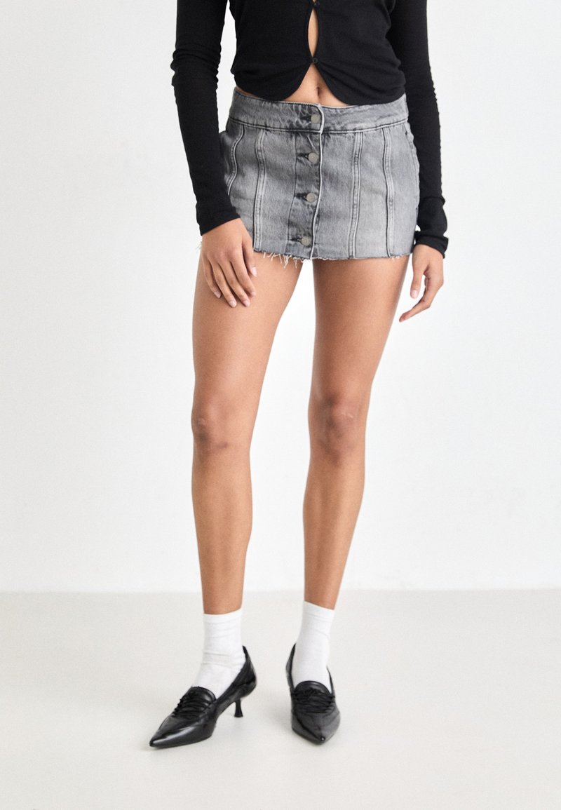Jupe en denim grise courte avec un ourlet effiloché, dotée de boutons sur le devant et de coutures verticales, associée à des chaussures noires et des chaussettes blanches.