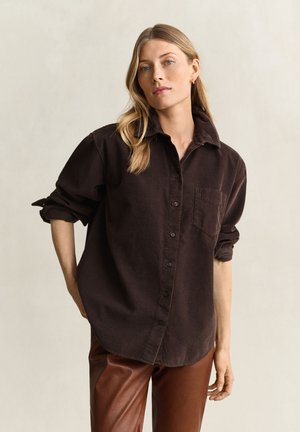 Femme aux longs cheveux blonds portant une chemise marron foncé boutonnée et un pantalon en cuir marron, debout devant un fond clair et uni.