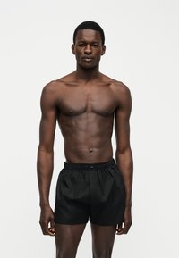 Μυώδης άνδρας με σκούρο δέρμα στέκεται μπροστά σε λευκό φόντο, φοράει μαύρα σορτς με ελαστική μέση και κοιτάζει μπροστά με ουδέτερη έκφραση.