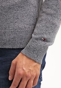 Pull gris en maille avec des poignets côtelés et un petit patch logo rouge, blanc et bleu. La texture est douce avec un motif moucheté.