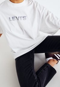 Camiseta blanca de algodón con mangas cortas, que presenta un logo gris de "LEVI'S". Lleva puesta con jeans oscuros y zapatillas, en una posición sentada.