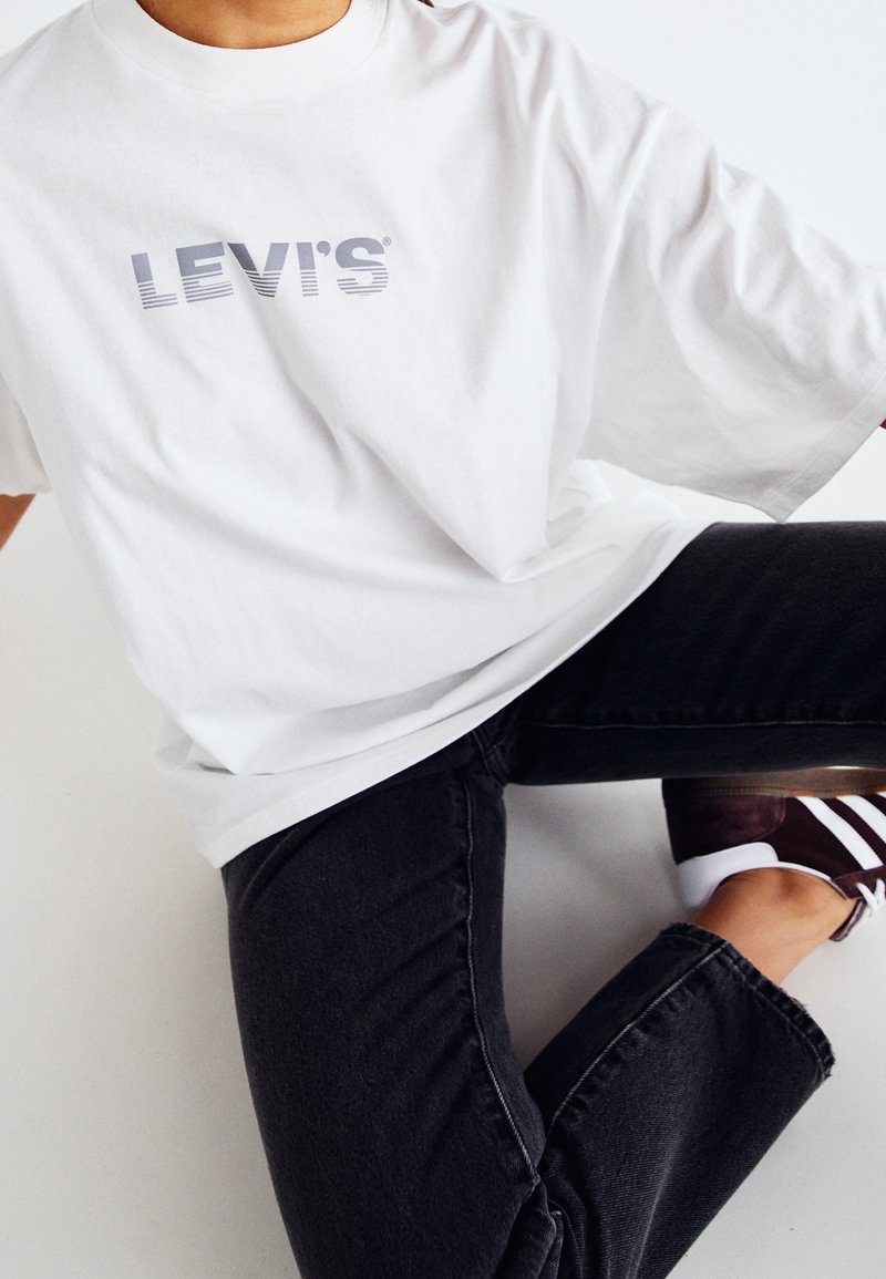 Camiseta blanca de algodón con mangas cortas, que presenta un logo gris de "LEVI'S". Lleva puesta con jeans oscuros y zapatillas, en una posición sentada.