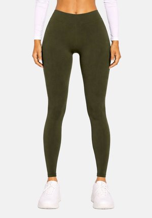 Femme portant des leggings taille haute vert olive et des baskets blanches, debout, les bras relâchés le long du corps devant un fond uni.