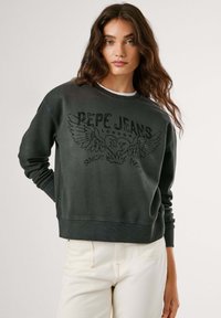 Sweatshirt grigio con scollatura a girocollo, caratterizzato da un design grafico nero con ali e testo, abbinato a jeans chiari. Texture morbida.