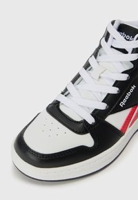 Tuvojuma attēls ar melnu, baltu un sarkanu Reebok sporta apavu ar baltām šņorēm un caurumiem purngalā uz baltas fona.