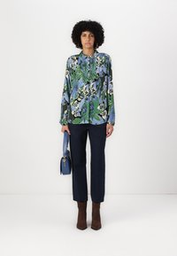 Long-sleeve blouse with a green, blue, and white floral design; paired with navy trousers and brown suede ankle boots; holding a blue handbag.
