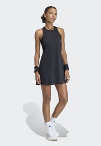 Frau trägt ein schwarzes ärmelloses Tennisdress, schwarze Handgelenkbänder, weiße Socken und weiße Turnschuhe, steht mit einem leicht gebeugten Bein.