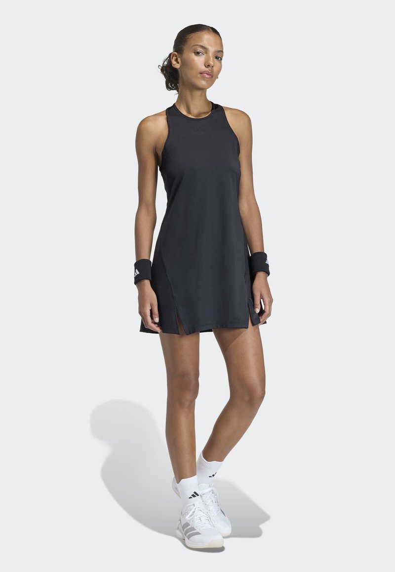 Frau trägt ein schwarzes ärmelloses Tennisdress, schwarze Handgelenkbänder, weiße Socken und weiße Turnschuhe, steht mit einem leicht gebeugten Bein.