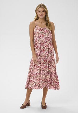 Kvinde iført en ærmeløs, pink og rød blomsterprint midi-kjole med tynde stropper og brune sko med lave blokhæle, stående på en hvid baggrund.