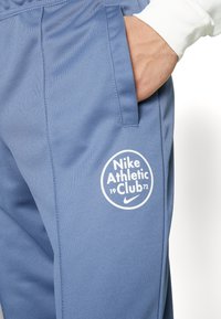Nike Sporthosen aus blauem Stoff, mit einem runden weißen Logo und dem Text "Nike Athletic Club 1972". Glatte Textur mit seitlichen Taschen.