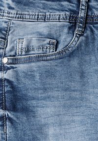 Džínové kalhoty v světle modré barvě s hladkou texturou, obsahující detail malého kapsičky, stříbrné kovové části a kontrastní žluté prošívání.