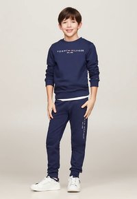 Completo felpa e pantaloni della tuta color blu navy, tessuto di cotone con logo bianco e rosso. Sneakers bianche, dettagli con polsini e cintura a costine.