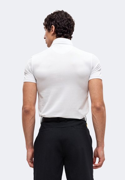 Homme aux cheveux bouclés foncés portant un polo blanc ajusté à manches courtes et un pantalon noir, de dos sur un fond clair uni.