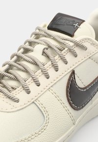 Nike sneakers med en cremet læderoverdel, brun swoosh og teksturerede accenter. Har beige snørebånd og et brunt logo-tag på tungen.