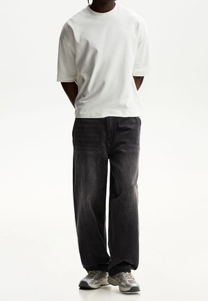 Osoba ubrana w prostą, białą oversize koszulkę, luźne czarne dżinsy i szare trampki, stojąca z rękami za plecami.