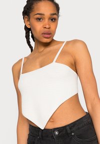 Top crop branco de malha com alças finas, apresentando uma bainha diagonal que se estende para uma forma pontiaguda. O tecido texturizado acrescenta interesse visual.