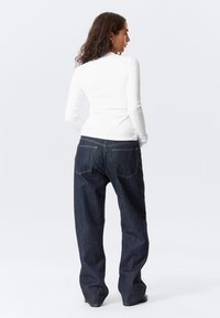 Haut blanc à manches longues et jean large bleu foncé. Le denim comporte deux poches arrière avec des coutures contrastées.