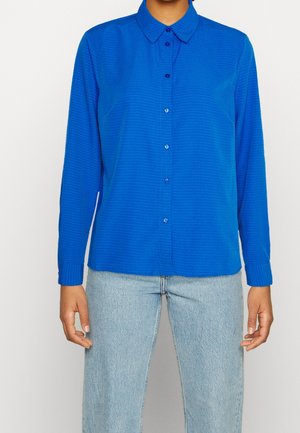 Persoon die een blauwe blouse met knopen, lange mouwen en kraag draagt en lichtblauwe spijkerbroek, staand tegen een effen achtergrond.