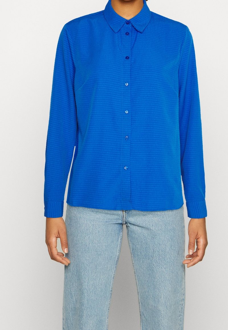 Personne portant une chemise bleue à manches longues boutonnée avec col et un jean en denim bleu clair, debout devant un fond uni.