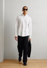 Camisa blanca de manga larga y botones, pantalones anchos negros, zapatos negros y un maletín negro. Las gafas de sol completan el look profesional.