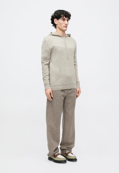 Beige hætte sweater med en frontlomme og snoretræk, parret med lysebrune bukser. Sættet afsluttes med grønne og sorte sko.