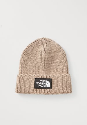 Beige gebreide beanie met opgevouwen rand en zwart rechthoekig logo-patch met de tekst "THE NORTH FACE" in witte letters.