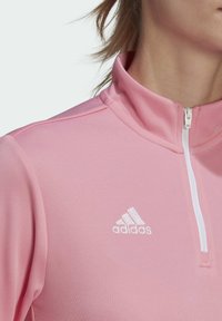 Light pink athletic half-zip top featuring a white zipper and embroidered logo. Material is smooth and breathable with a textured finish.