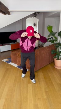 Červená baseballová čiapka, fialovo-ružová vzorovaná bunda na zips, čierne nohavice a strieborné topánky, vnútorné prostredie s kvetináčom v pozadí.