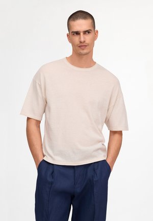 Mand med kort hår iført en beige oversized kortærmet t-shirt og mørkeblå bukser, står med hænderne i lommerne og kigger til siden.