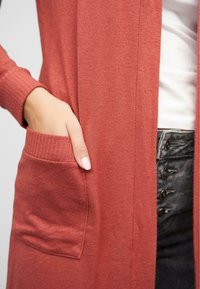 Lang, rustfarget cardigan laget av mykt stoff, med ribbede mansjetter og en lomme på venstre side. Glatt tekstur med et uformelt design.