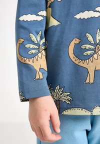 Pitkähihaine sininen paita, jossa on dinosaurusten ja palmun kuvio. Pehmeä puuvillakangas, näkyvä ompelu ja rento leikkaus; yhdistetty vaaleansinisiin housuihin.