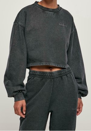 Osoba w ciemnoszarej krótkiej bluzie i pasujących wysokich dresach, odsłaniająca brzuch i stojąca na jednolitym tle.