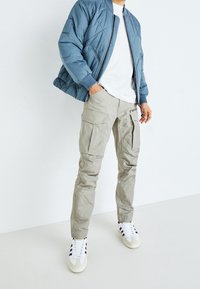 Modrá prošívaná bomber bunda, bílý tričko, béžové cargo kalhoty s kapsami a bílé tenisky s černými pruhy. Důraz na funkčnost.