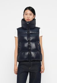 Navyblauwe puffer vest, glanzend materiaal, hoge kraag, ritssluiting en zijzakken met drukknoopjes. Draagt over een wit shirt.