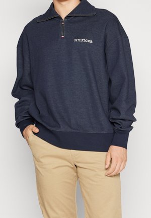 Osoba nosząca granatową bluzę z suwakiem do połowy z logo "HILFIGER" na piersi, w połączeniu z beżowymi spodniami, jedną ręką w kieszeni.