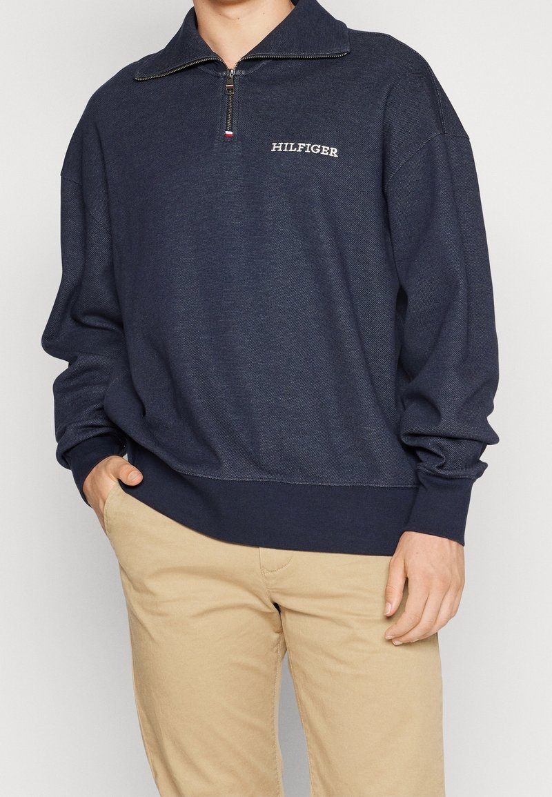 Personne portant un sweat-shirt bleu marine à fermeture éclair quart avec le logo "HILFIGER" sur la poitrine, associé à un pantalon beige, une main dans la poche.
