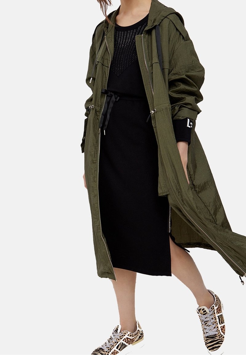 Femme portant un long manteau zippé vert olive, une robe midi noire et des baskets à imprimé animalier mêlant motifs zèbre et léopard.