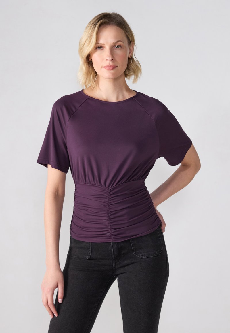 Lilla kortermet topp med samlet midje, laget av elastisk stoff. Jevn tekstur, rund halslinje og raglanermer. Sammen med svarte jeans.