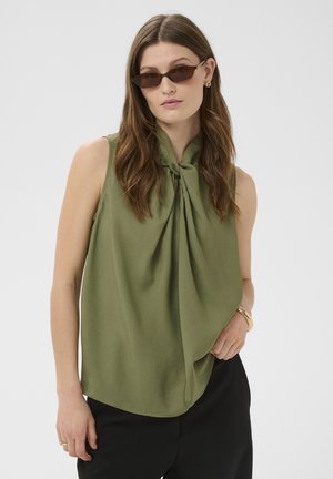 Vrouw draagt een olijfgroene mouwloze blouse met een grote knoop aan de voorkant, zwarte broek, cat-eye zonnebril en gouden sieraden, poserend tegen een grijze achtergrond.