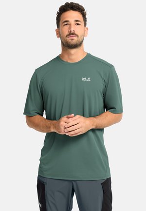 Uomo con capelli corti e ricci che indossa una maglietta verde a maniche corte Jack Wolfskin e pantaloni cargo grigi, in piedi con le mani incrociate.